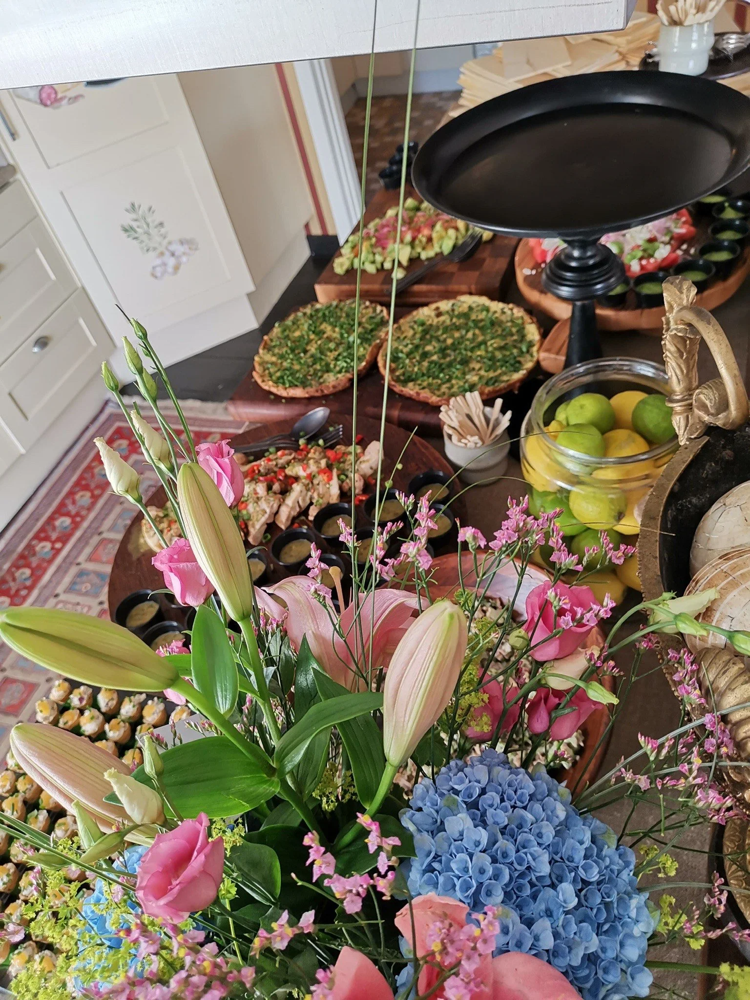 Blommig arrangemang med rosa liljor, blå hortensia och andra blommor framför ett bord med tårtor, bakverk och frukt.