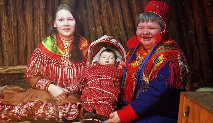 Bathed in warm light and surrounded by wooden walls, three generations of Sámi women gather in a moment of joy and tradition. At the center rests a sleeping baby, safely swaddled in a crimson giehtkka — the traditional Sámi cradleboard — laced with p