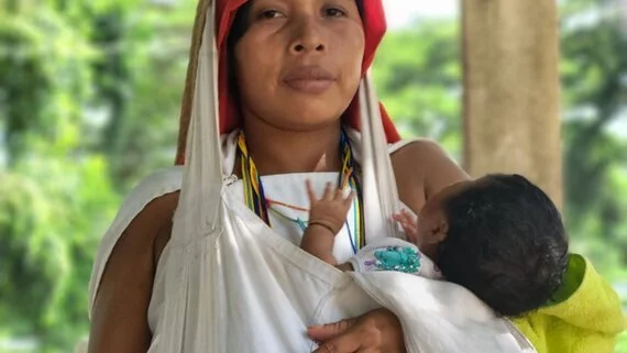 A Wayuu mother carries her baby close in a traditional cloth rebozo, cradled against her heart. Her striking head covering and beadwork speak of cultural identity and ancestral pride, while her calm strength fills the frame. Among the Wayuu people of