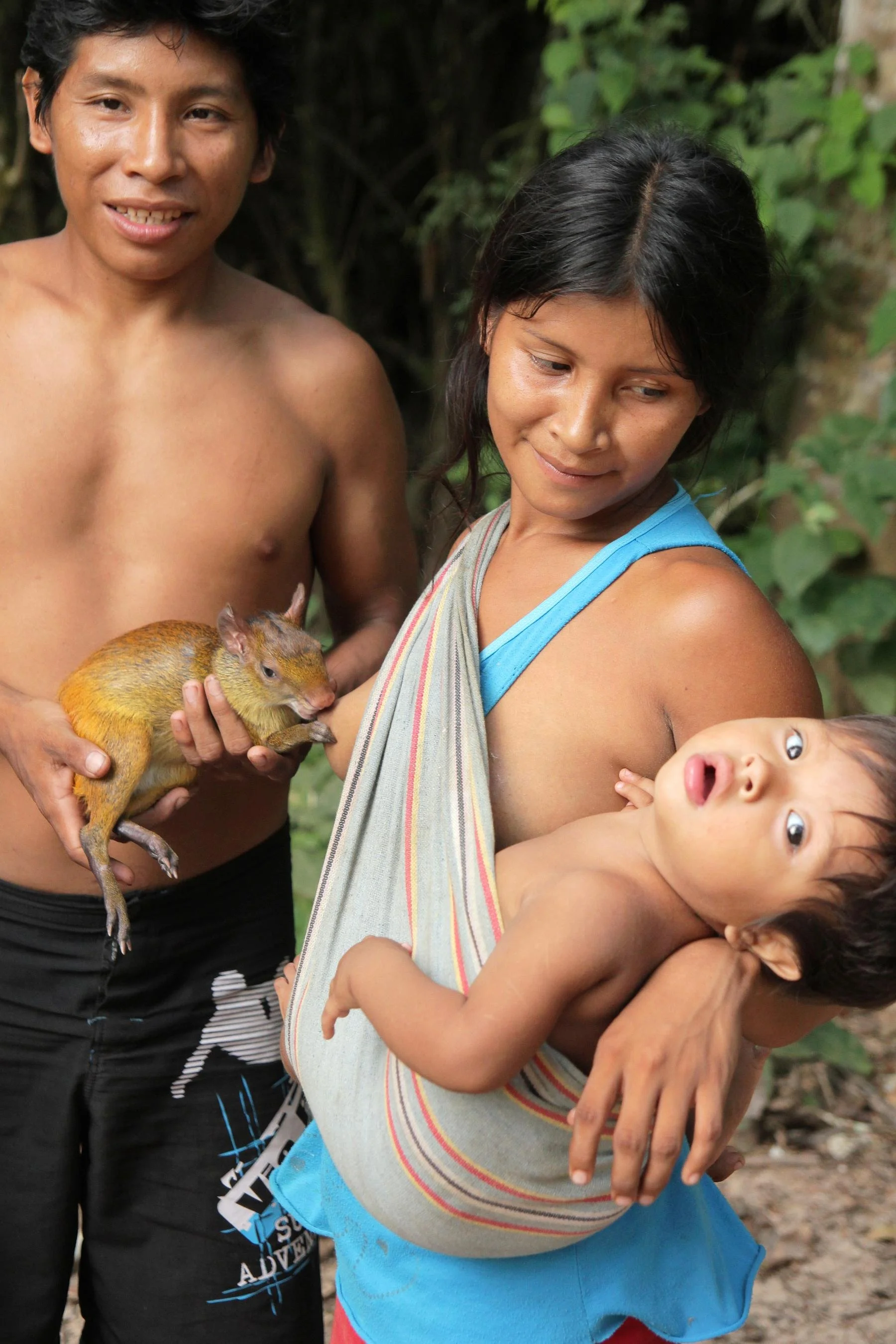 This image shows an Indigenous family from the Peruvian Amazon. The mother is carrying her child in a traditional cloth sling across her shoulder — a common babywearing practice among many communities in the region. The style of the sling and the for