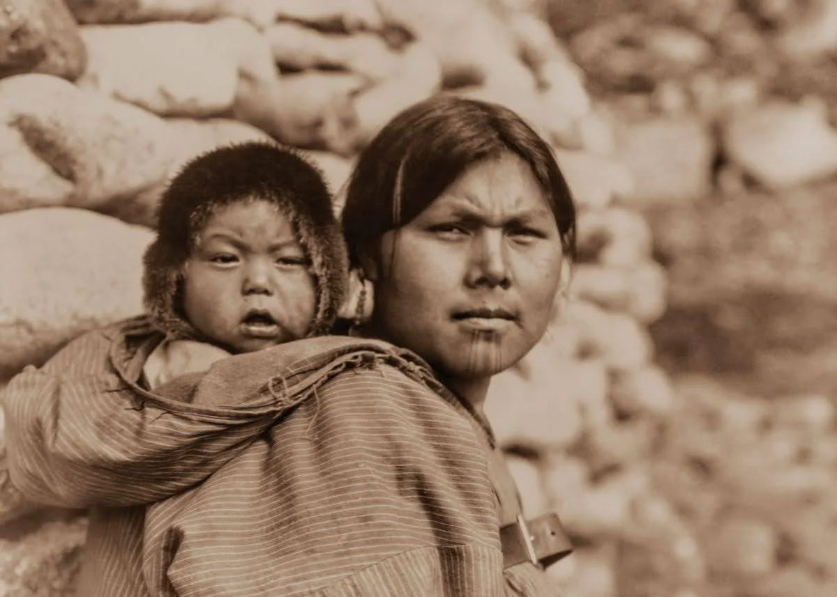 In this striking historical portrait, a mother from Little Diomede Island, Alaska, carries her child high on her back. The child, bundled warmly in a fur-lined hood, peers out with a calm, watchful gaze. The mother’s face is steady and strong, adorne