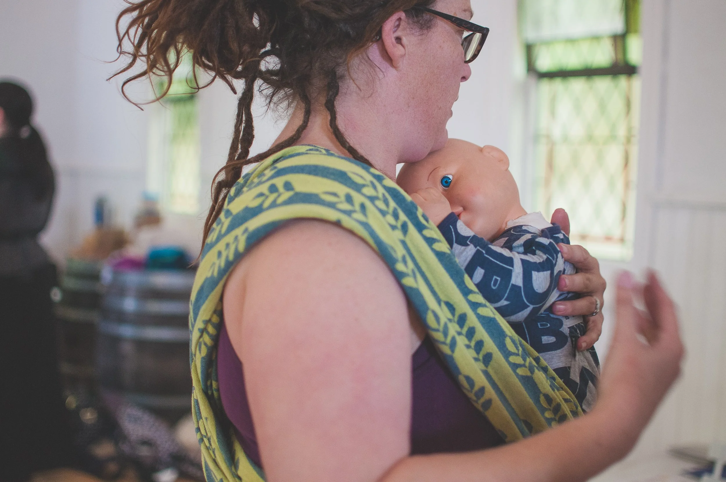 A woman with curly hair and glasses holding a baby close to her chest inside a room with windows and natural light.