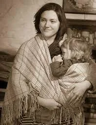 A modern mother smiles softly as she holds her toddler close, wrapped securely in a traditional woollen shawl. The cloth is drawn across her body in the classic Welsh shawl carry — a method rooted in centuries of caregiving, adapted here with warmth