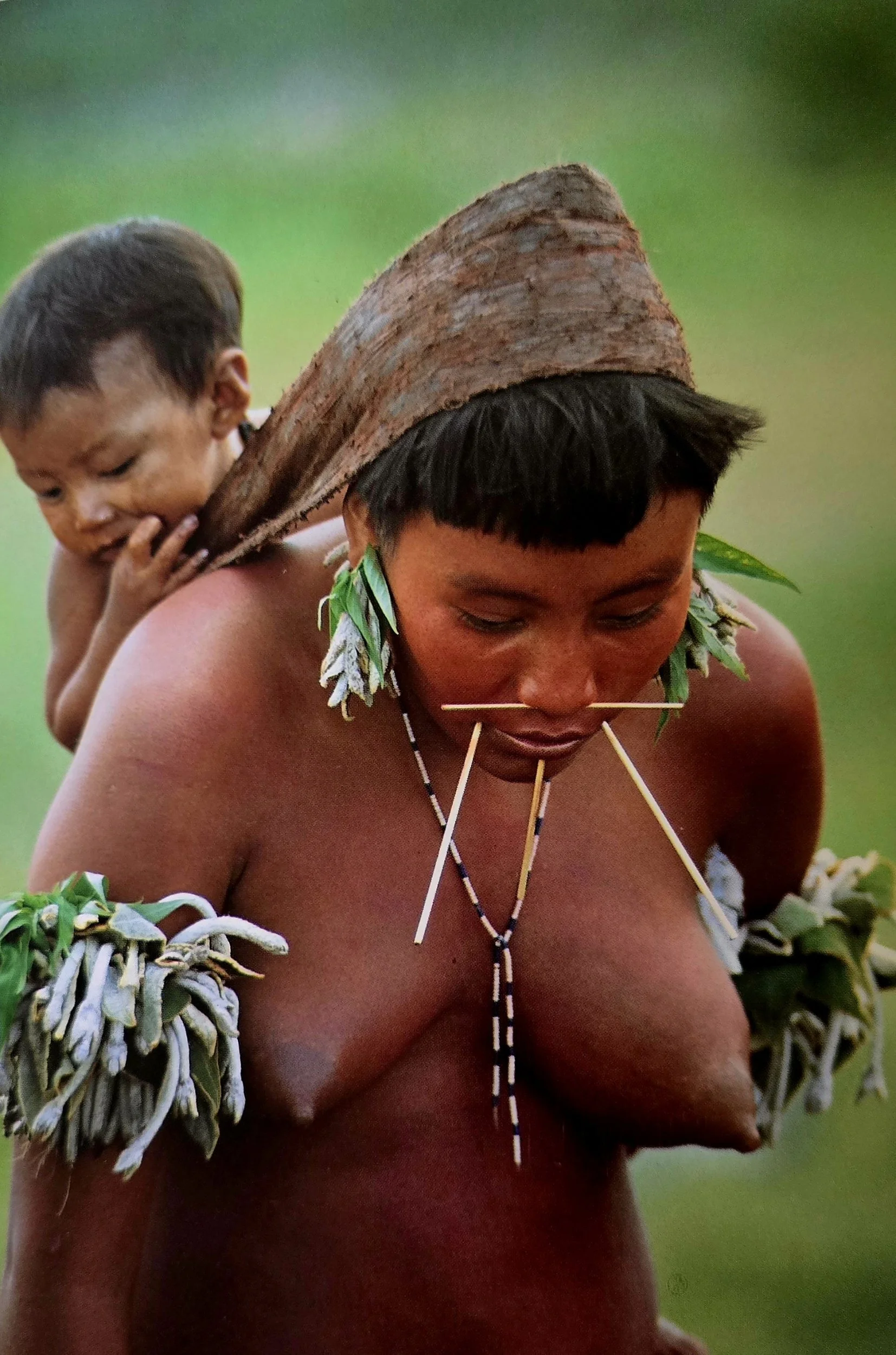 Against a blurred wash of green forest, a mother walks forward with steady focus, her young child nestled high on her back. The baby is held securely beneath a folded bark cloth, an instinctive sling that needs no buckles or seams—just the knowing ha