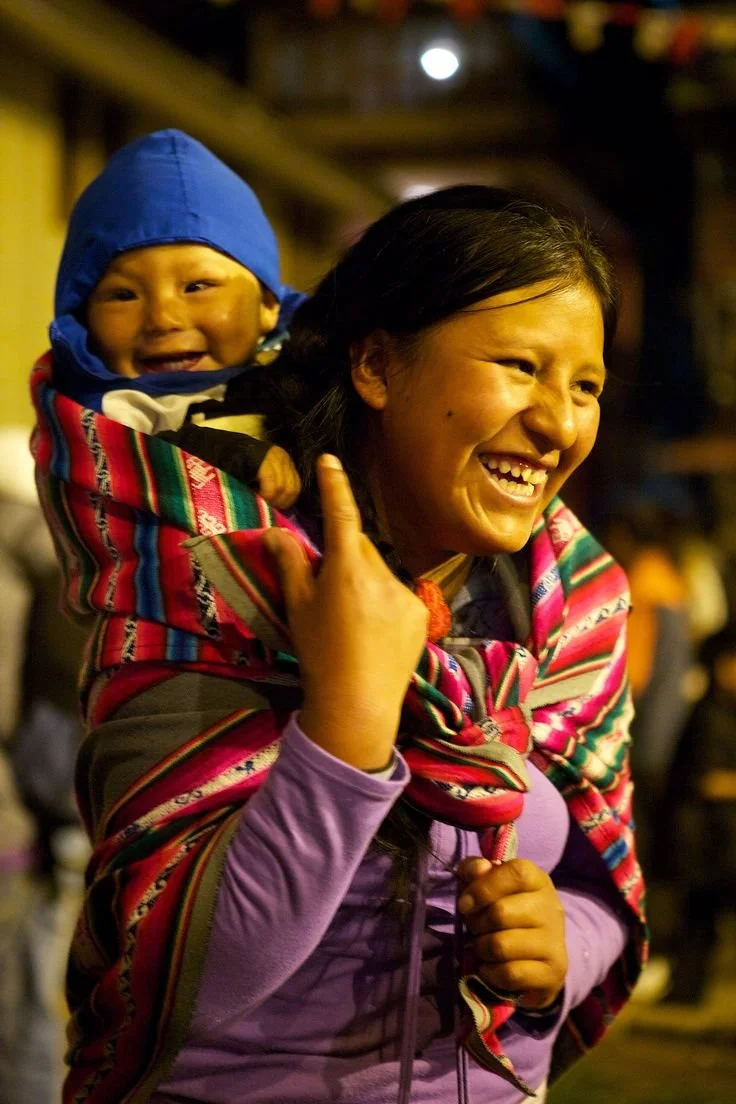 In the golden warmth of evening light, a mother laughs with her whole face, eyes crinkling as joy spills over. Wrapped snugly against her back, her baby grins just as wide, peeking out from a vivid aguayo cloth tied with skill and care. The bold stri