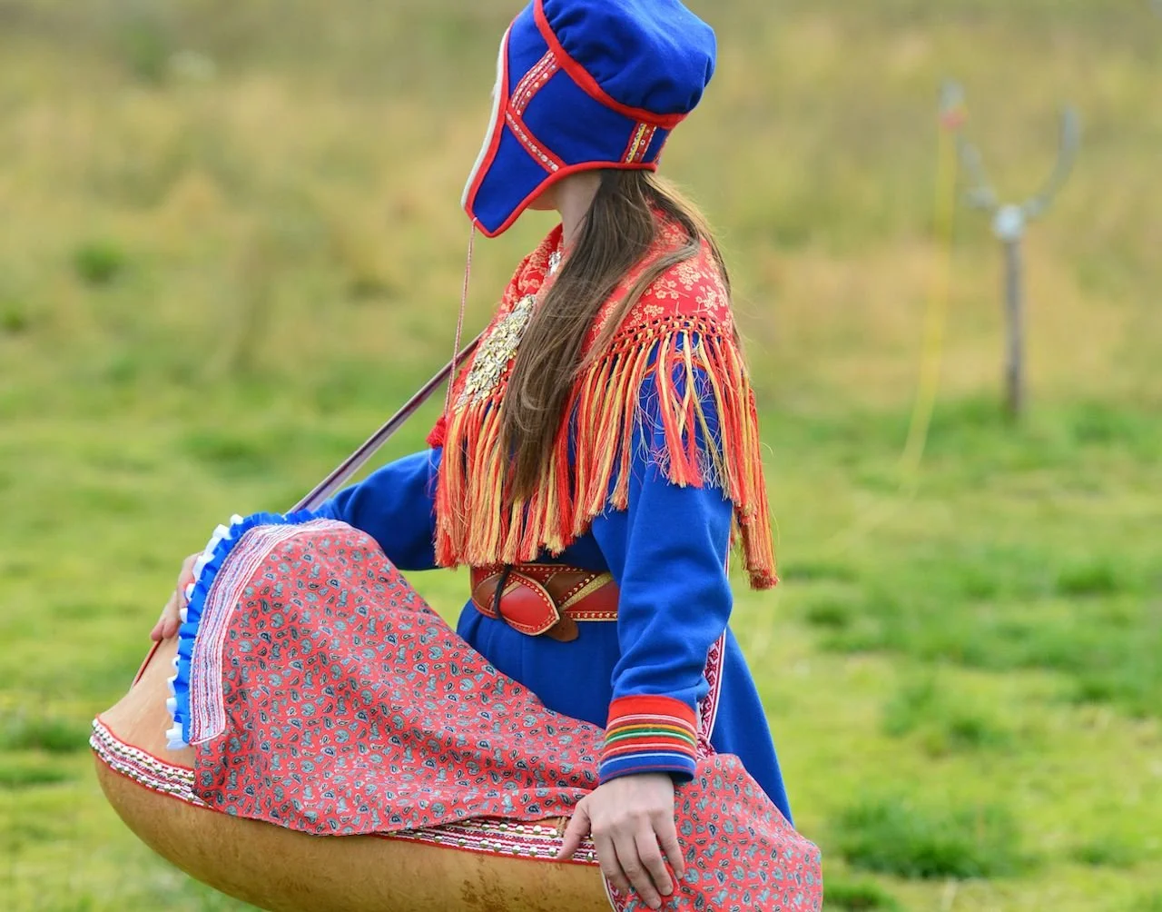 In the rolling green fields of Sápmi, a Sámi woman wears the richly embroidered gákti — traditional Sámi dress — and carries her baby in a beautifully crafted komsekule, a cradleboard known for its protective and spiritual significance. The cradle, o