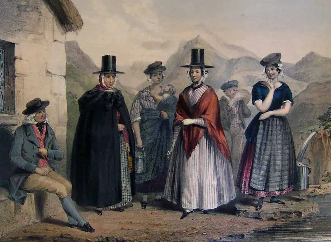 In a quiet Welsh village nestled among the mountains, a group of women gather in conversation, their clothing capturing the elegance of 19th-century rural life. Two wear the iconic tall black Welsh hats, paired with long shawls and layered skirts. On