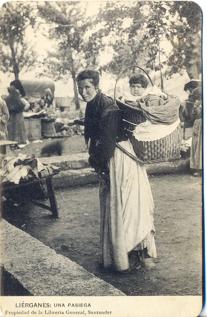 In the market town of Liérganes, in the Cantabria region of northern Spain, a Pasiega woman carries her child in a large basket strapped across her back. This historic photograph offers a rare glimpse into the daily rhythms of life among the Pasiegos