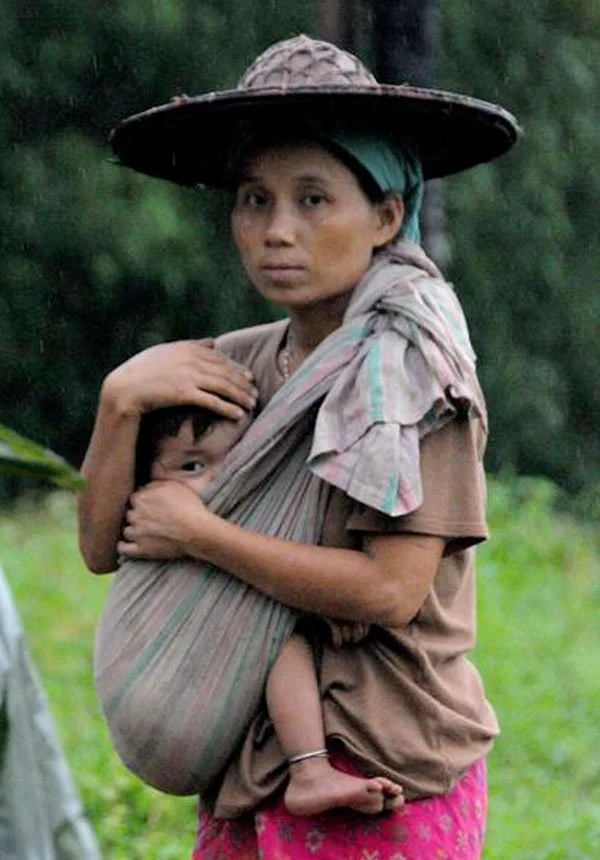 A mother stands quietly in the rain, her expression calm and steady beneath the wide brim of a traditional woven hat. She carries her child in a simple piece of cloth, knotted over one shoulder and cradling the baby snugly against her chest. The carr