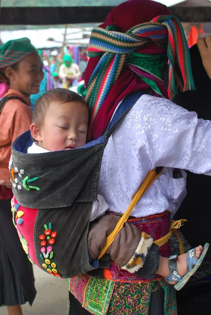 A small child sleeps soundly in a traditional back carrier, surrounded by the colour and texture of highland life. The caregiver, dressed in the vibrant embroidered clothing of the Flower Hmong people of northern Vietnam, moves through the bustle of