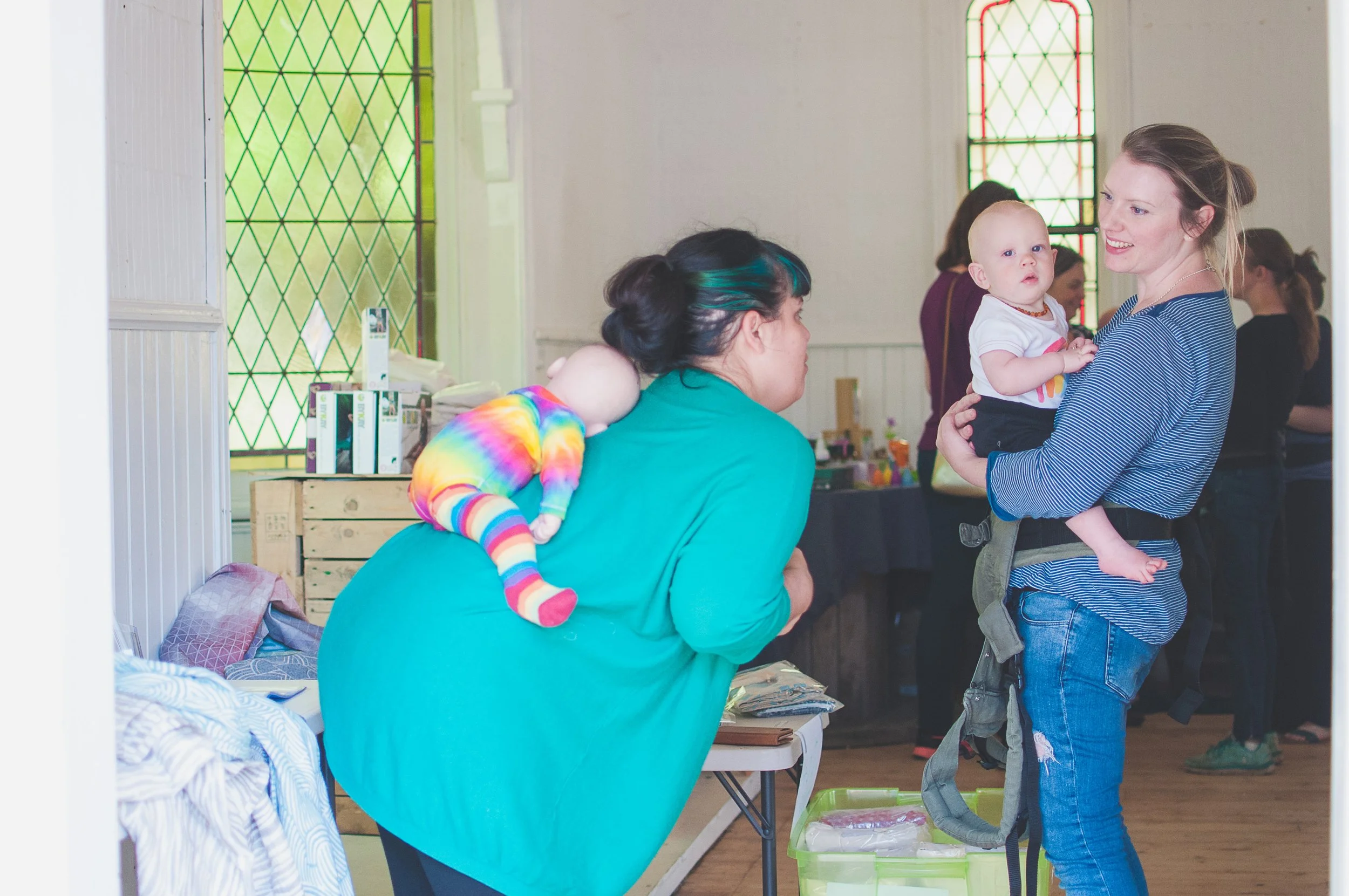 A woman in a teal top bends over a table to speak with a blonde woman who is holding a baby. The blonde woman is smiling, wearing a blue striped shirt and jeans. The baby is dressed in white with colorful designs. The scene appears to be indoors with stained glass windows in the background.