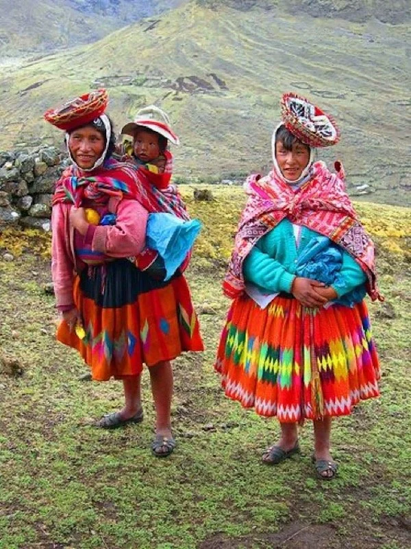 High in the Peruvian Andes, two Quechua women stand side by side in a mountain clearing, wrapped in the warmth of tradition and colour. Their full, handwoven polleras ripple in bold reds, oranges, and zigzagged rainbows, echoing the vibrant land they