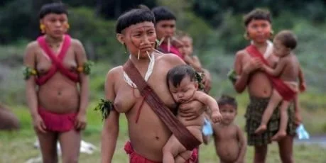 Amidst an open clearing, a group of Yanomami women gather — their bodies painted with tradition, their adornments telling stories of belonging. At the center, one mother walks with quiet strength, her baby cradled in a broad woven sling slung diagona