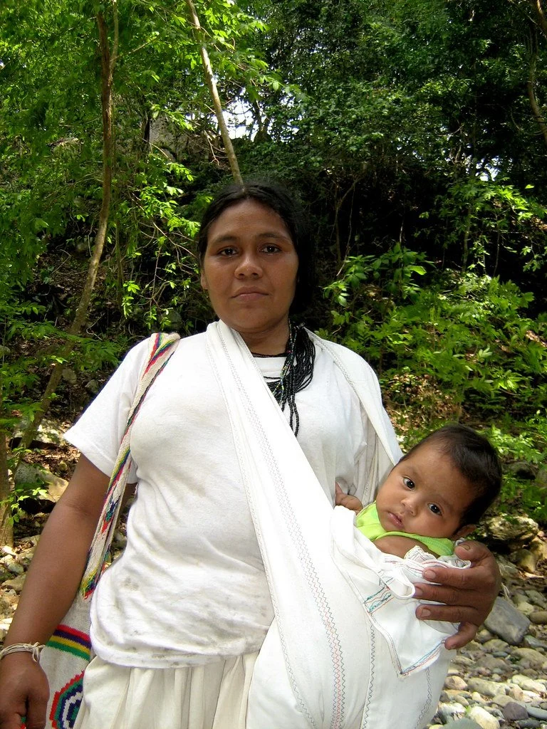 A Kogi mother stands quietly with her baby nestled in a handwoven sling, the fabric gathered over one shoulder in a wide supportive carry. Her white garments reflect the Kogi people's deep connection to the spiritual purity of nature — a tradition ro