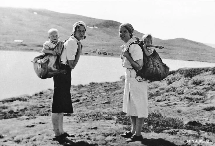 Two Norwegian mothers pause on a hillside above a mountain lake, each carrying a child securely in a large woven pack basket. Their clothing — calf-length skirts, practical blouses, and kerchiefs tied around their heads — suggests a 1930s or 1940s ru