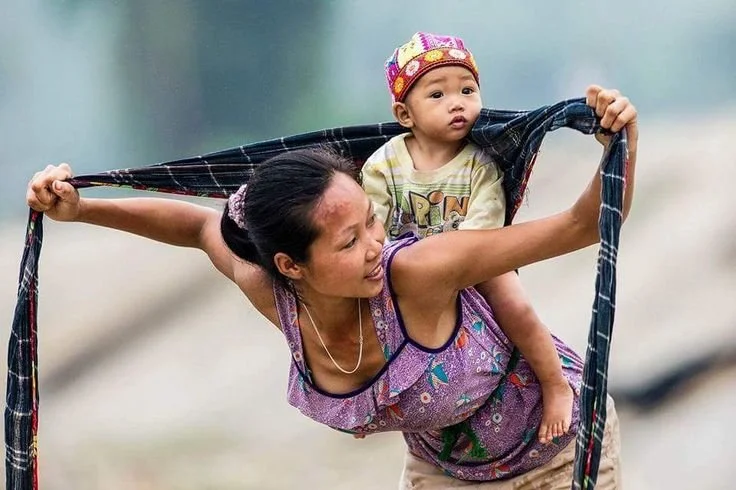  In this beautiful moment, a young woman from Southeast Asia — likely Vietnam or a nearby hill tribe region — expertly ties her baby onto her back using a long, simple woven cloth. We see her leaning forward, smiling, as she gathers and tightens the 