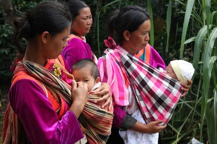  In this vibrant image, three Kayan women from Myanmar (Burma) wear their babies close in traditional shoulder cloths. These carriers, likely woven on a backstrap loom, are tied securely across one shoulder in a style that functions similarly to a mo