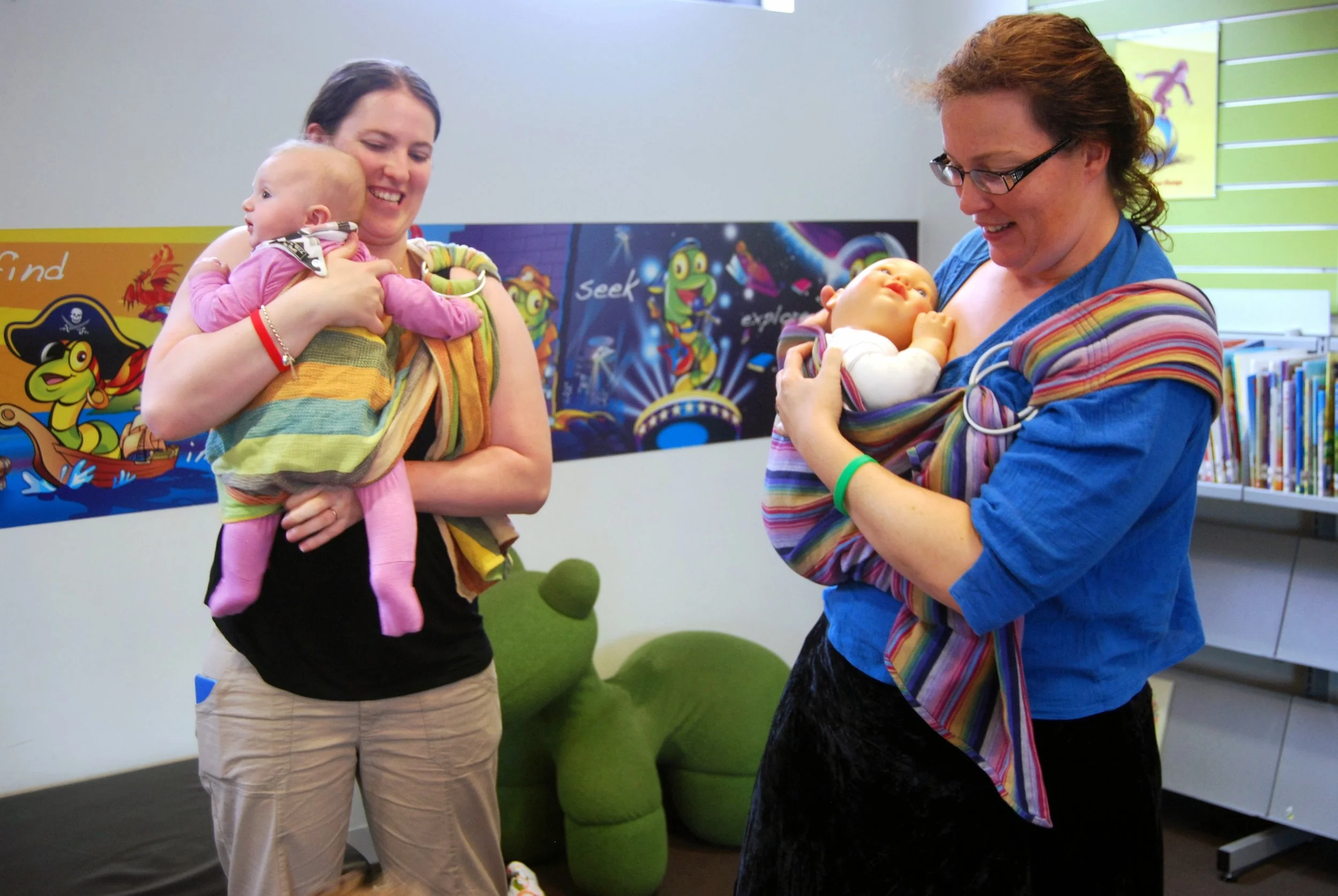  In a cheerful children’s library corner in Australia, two women share a moment of learning and laughter as they explore the art of babywearing with ring slings. One holds her baby snug in a rainbow-striped wrap, adjusting the fabric through the slin