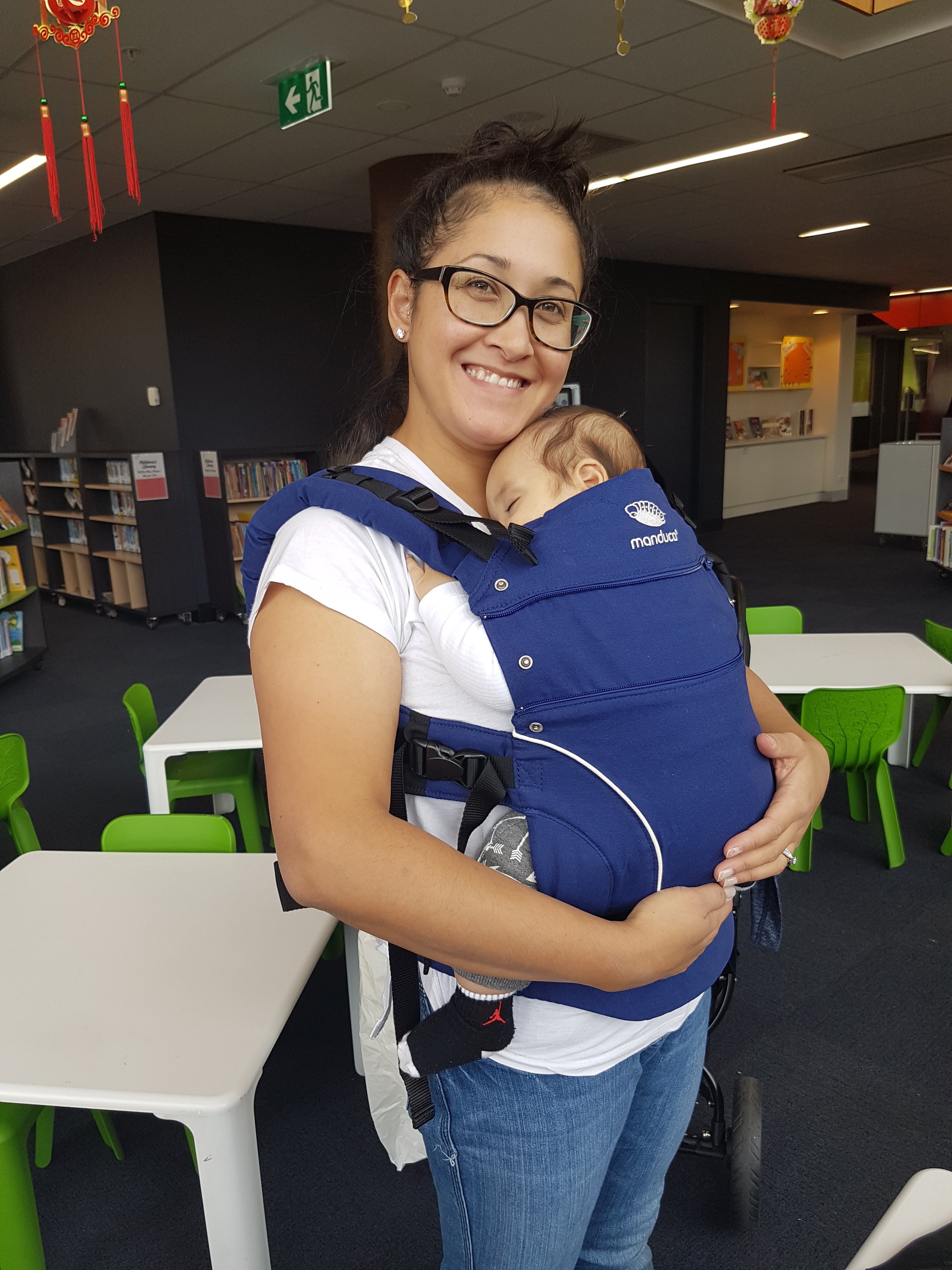  A mother beams as she holds her sleeping baby close in a structured soft carrier — the Manduca, a popular ergonomic buckle carrier inspired by the traditional  meh dai . The baby rests snugly in an upright position, legs well supported, and head tuc