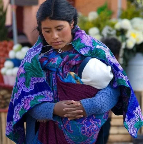  A young Tzotzil Maya mother from the highlands of Chiapas, Mexico, carries her baby snugly against her chest using a rebozo—an intricately woven rectangular cloth traditionally used throughout Mesoamerica. The baby is nestled securely within deep fo