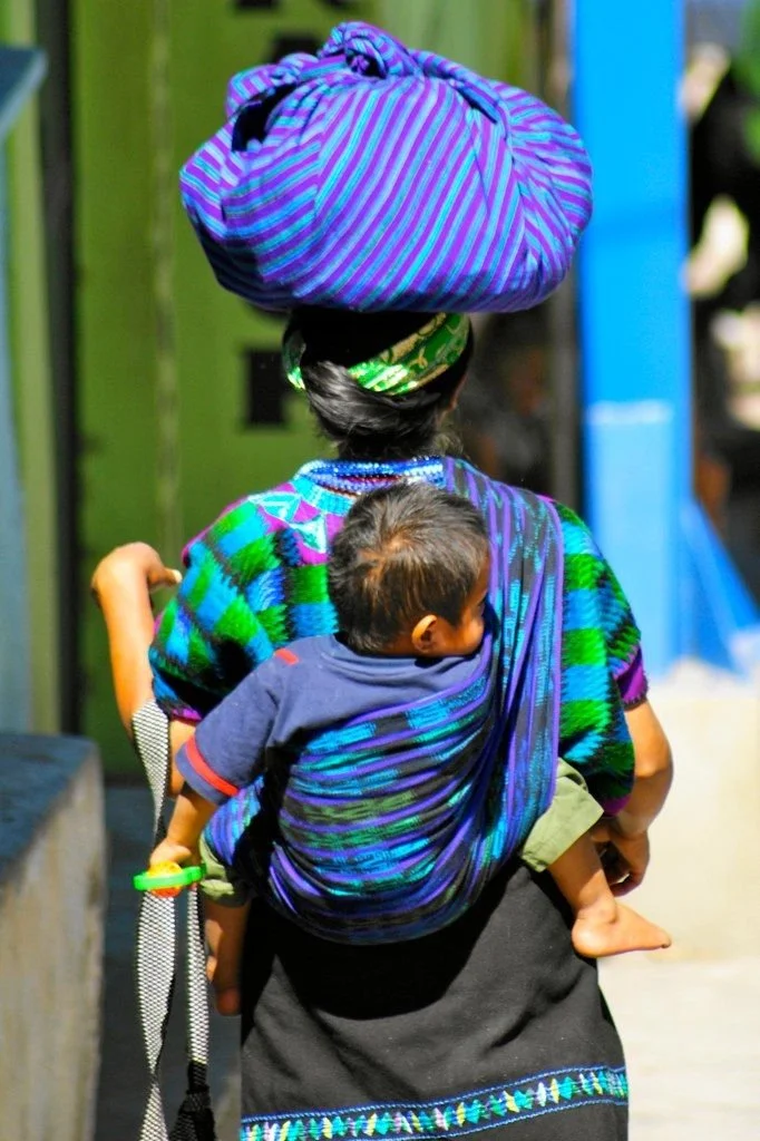  A K’iche’ Maya woman from the highlands of Guatemala carries her toddler on her back using a traditional handwoven  rebozo , wrapped securely around them both and tied over one shoulder. The vivid blue, green, and violet threads of the cloth match h