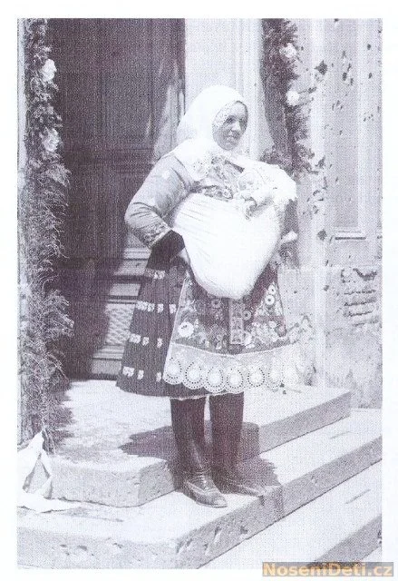 A mother stands on stone steps outside a grand doorway, her head wrapped in a white scarf and her embroidered blouse bright beneath a tailored vest. The wrap around her waist swells with the bundled shape of her baby, who is tucked into a structured