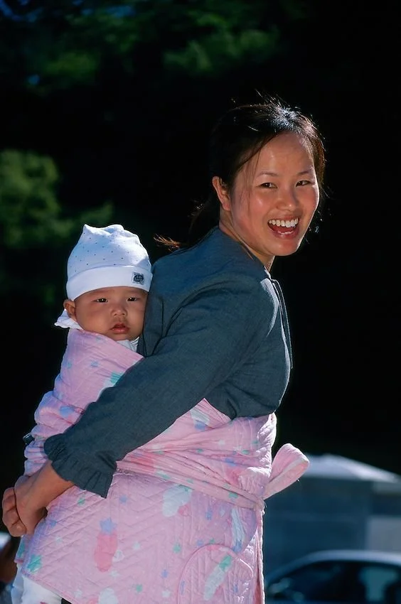 A mother smiles brightly under the midday sun, her body angled in gentle movement, her baby nestled snugly against her back in a thickly padded, pastel-pink blanket wrap. The carrier is a modern adaptation of the podaegi, a traditional Korean blanket
