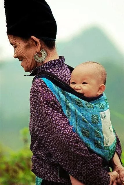 A beaming baby is carried high on the back of an older woman, secured in a richly woven turquoise and indigo cloth carrier. The baby’s eyes sparkle with joy, framed by the embroidered panels of the traditional textile that wraps snugly around their b