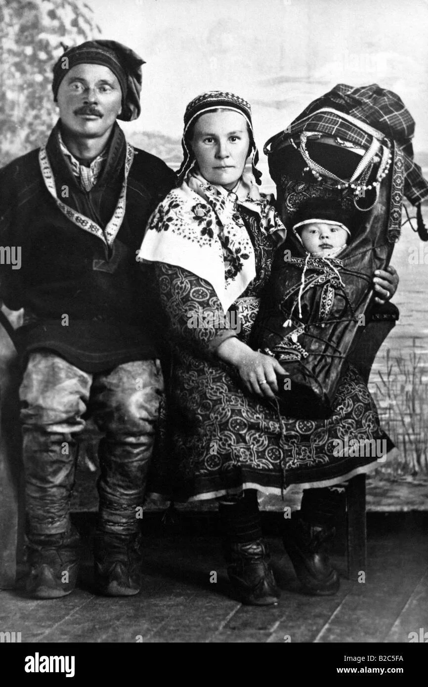 This striking studio portrait captures a Sámi family in traditional dress, with their baby nestled securely in a richly decorated komsekule — the distinctive Sámi cradleboard. The baby is wrapped and fastened with precision, held in place by intricat