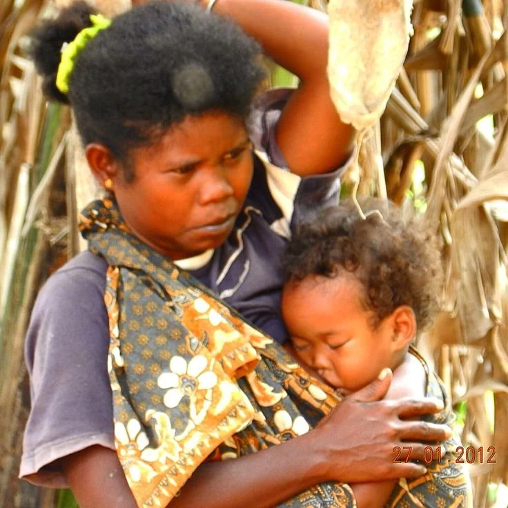 A mother draws her baby close, supporting a deeply sleeping child with one hand while the other adjusts a patterned cloth wrap across her shoulder. The child is held snugly in a front carry, with the fabric forming a gathered sling that supports the