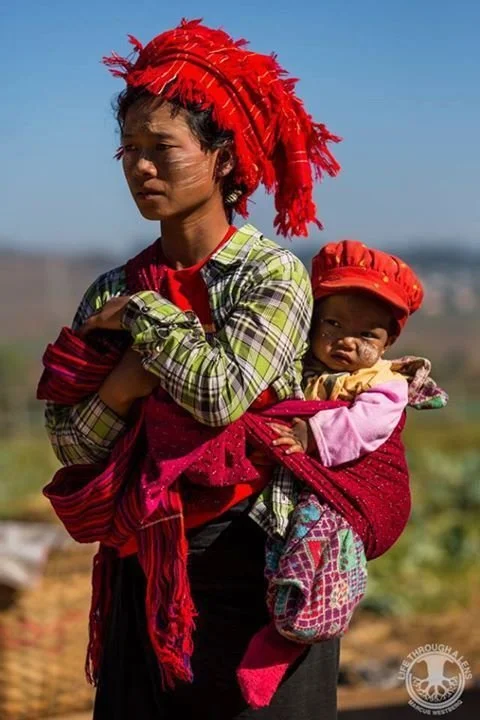 A mother stands beneath the bright sky, her posture calm and self-contained, as her child rests securely at her side in a deep red cloth wrap. Both are adorned with vivid red head coverings — hers a striking headdress of tasselled layers, the baby’s