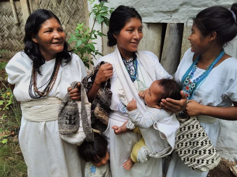 Three Arhuaco women share a moment of laughter, their white garments and bead necklaces catching the light outside a traditional dwelling. One mother nurses her baby in a sling, the cloth wrapping diagonally across her body to support the infant in a