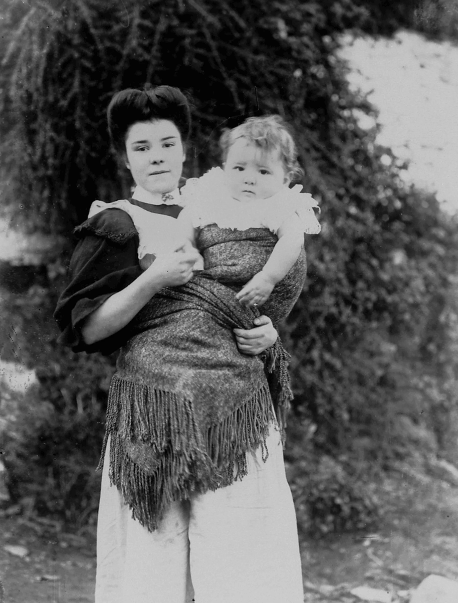 A young woman carries her child securely in a woollen shawl, the thick, fringed fabric wrapped snugly around their bodies and knotted at her back. Her high-necked blouse and upswept hair suggest the early 20th century — a time when fashion and functi