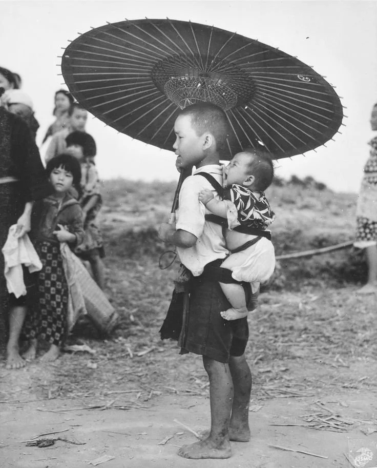 In this striking black-and-white photograph taken during or shortly after World War II, a barefoot boy stands tall beneath a large paper parasol, his younger sibling nestled securely on his back. The baby is carried high, upright, and close in a trad