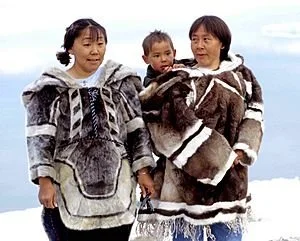 Two Inuit women stand side by side in traditional fur parkas, designed for the deep cold of the Arctic. One carries a child securely tucked into the amauti — a distinctive coat with a spacious hood used for babywearing. The child peers calmly over th