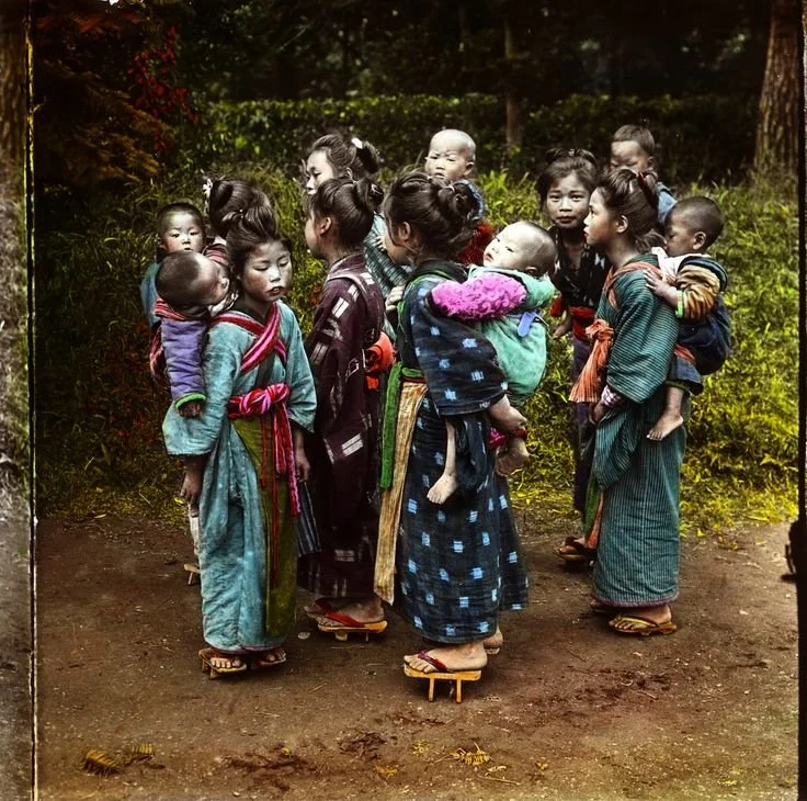 A group of young girls gathers in a forest clearing, their traditional Japanese clothing vibrant with indigo, crimson, and teal. Their hair is neatly tied, some with ribbons, others with simple knots, and almost every one of them carries a baby or to