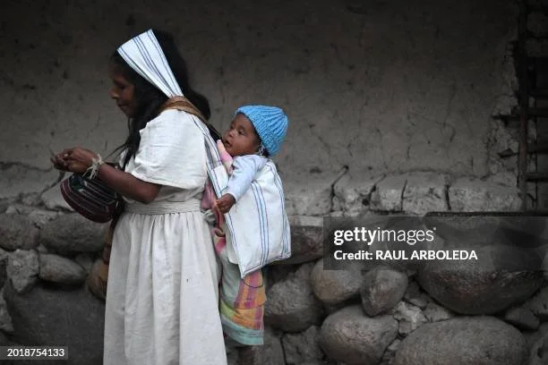 A young Kogi mother walks with purpose, her baby secured in a soft back carry with layers of woven cloth. The infant, dressed warmly in a blue knit cap and colourful wrap, rests quietly against her back—alert to the world, yet held close in safety. T