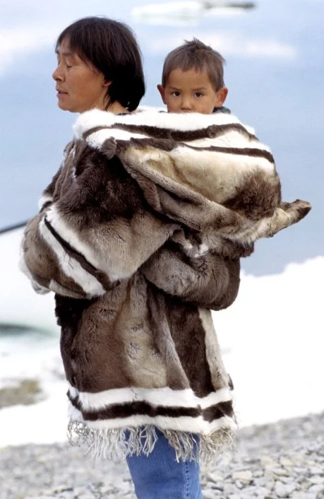 Against a backdrop of snow and sea, a mother carries her child nestled within a traditional Inuit amauti, expertly crafted from sealskin and trimmed with fur. The coat’s generous hood encircles them both, designed to accommodate the child in a pouch