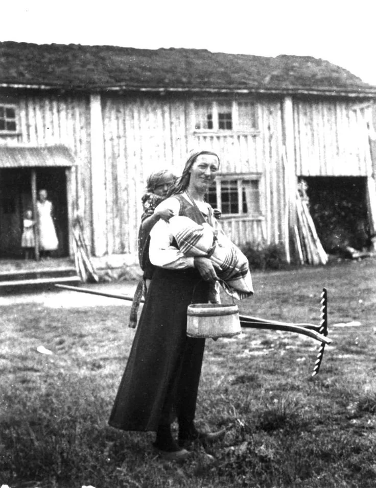 Outside a timber farmhouse, a woman smiles toward the camera, standing tall and balanced despite her load: a baby tied snugly to her back with a woven cloth, a large wooden rake resting over her shoulder, and a metal pail carried in one hand. Her sta