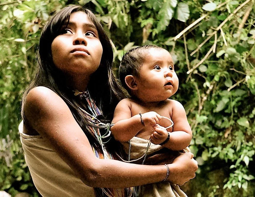 Bathed in soft jungle light, an older Kogi sister cradles her baby sibling in her arms. Her wrap dress, beaded necklaces, and calm focus reflect her early role as caregiver within the Sierra Nevada’s matrilineal rhythms. The baby, swaddled in a simpl