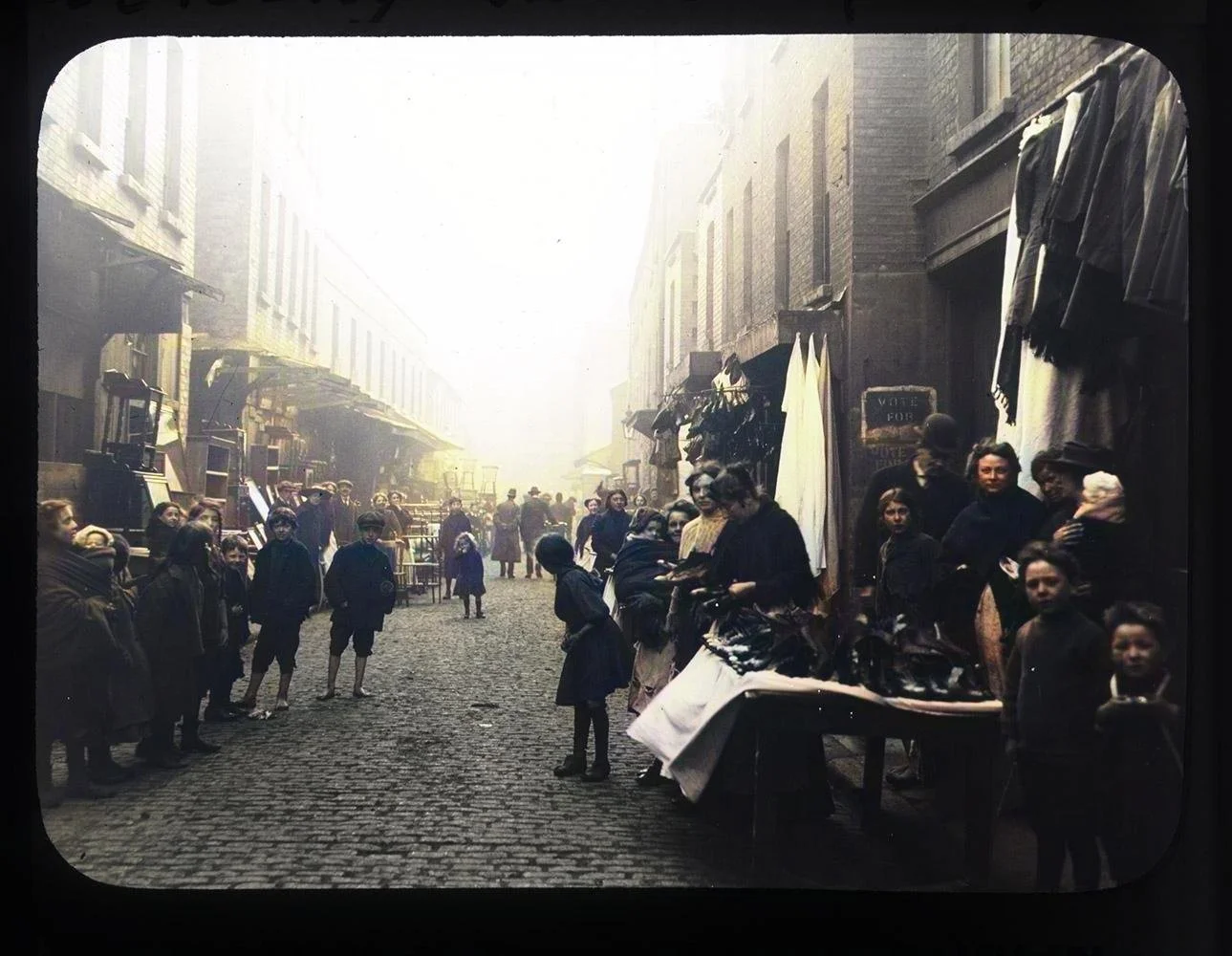 A narrow market street hums with life in early 20th-century Ireland. The cobbled lane is lined with traders and townsfolk, children gathered in small clusters, and tables piled with dark leafy produce. Long bolts of fabric hang from doorways, and a t