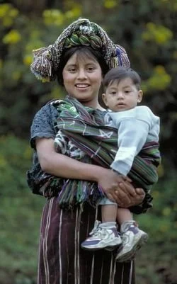 In this image, a Maya woman from Guatemala carries her child securely on her hip with the help of a woven rebozo or tzute, a cloth rich in both beauty and meaning. Her traditional huipil blouse and intricately wrapped headpiece proudly display her co