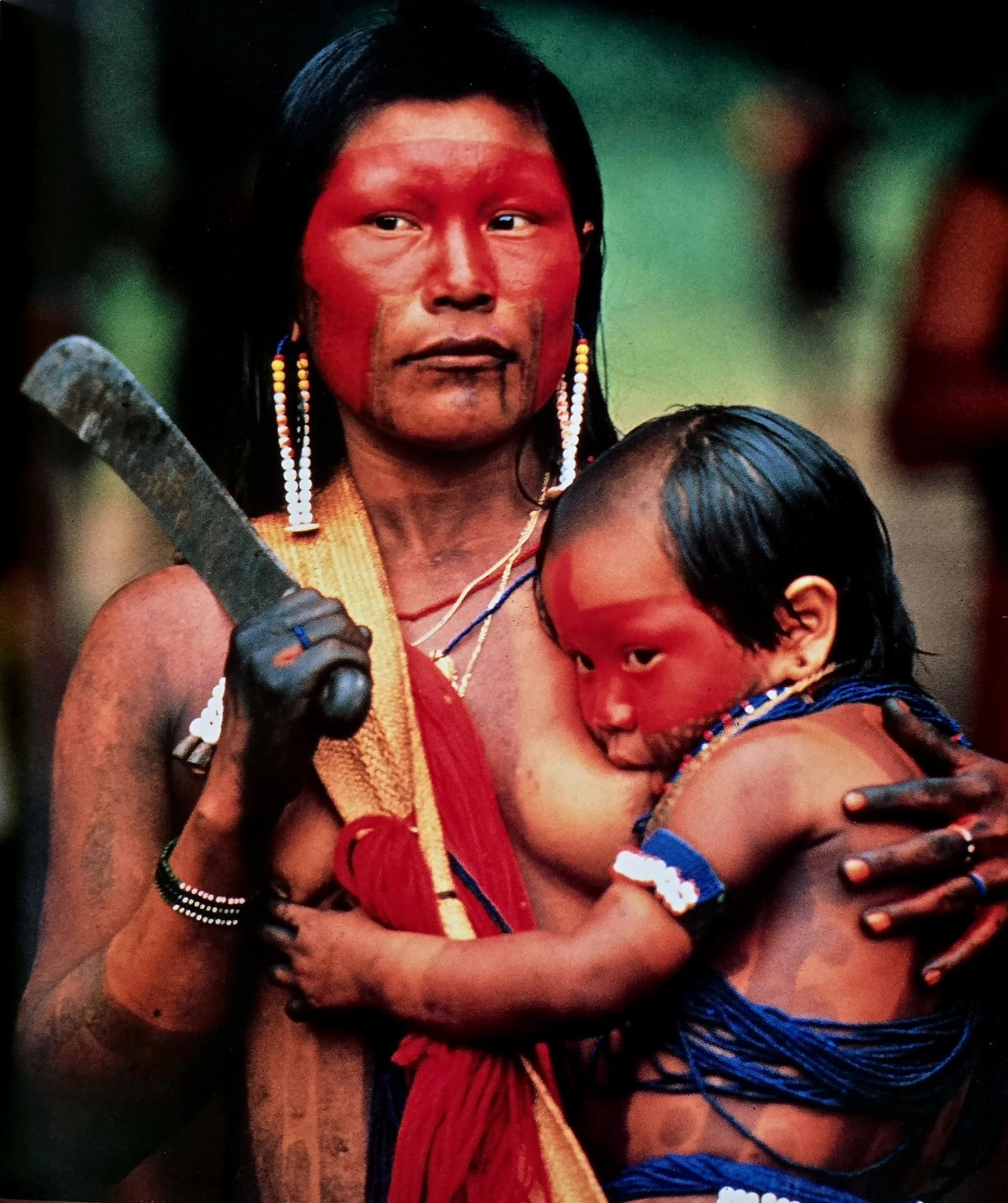 A Kayapo mother stands tall and grounded, her face and upper body vividly painted in sacred red—a colour drawn from the urucum seed, symbolising strength, life, and spiritual identity. Bright beaded earrings hang beside her face, and her long black h