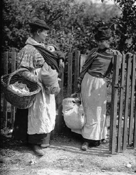 Two working-class women pause in conversation at a wooden gate in this early 20th-century photograph, dated around 1910. One carries a large wicker laundry basket on her hip, and the other, a heavy sack — each woman balancing the physical burdens of