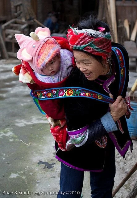 A woman bends slightly at the waist as she moves through an outdoor courtyard, her baby securely carried on her back in a richly embroidered textile carrier. Her posture is strong and practiced, one hand resting across her lower back in a familiar ge