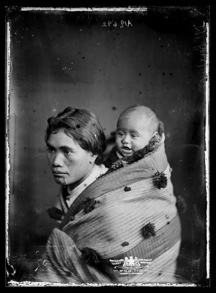 Wrapped in a beautifully adorned cloak, a Māori mother carries her baby high on her back, their closeness woven into the fabric of the garment and the moment. The korowai is decorated with tassels that signal both protection and cultural significance