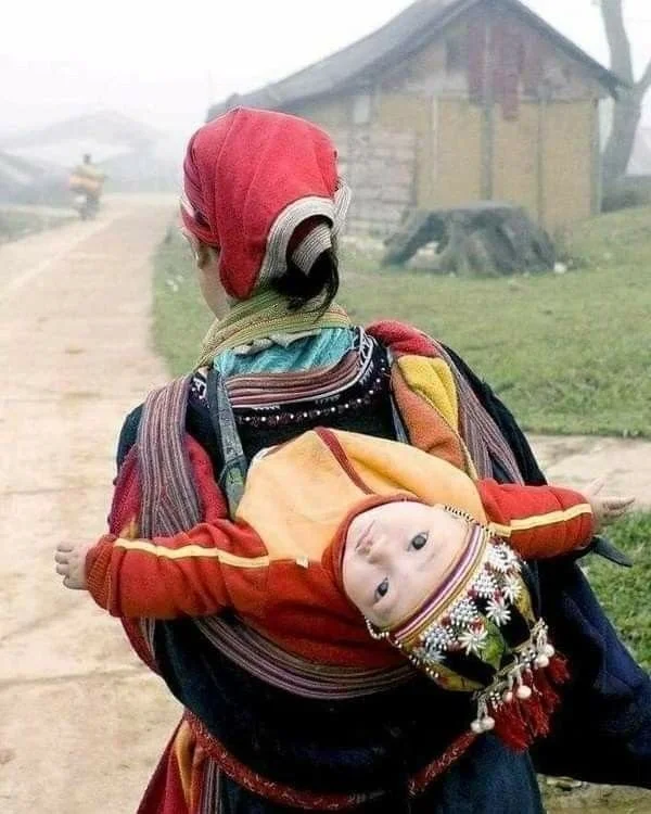 Wrapped in vibrant red and gold, a baby looks directly over their mother’s shoulder, body arched and arms outstretched toward the viewer. Their wide gaze and outstretched posture suggest alert curiosity — or perhaps the momentary strain of twisting t