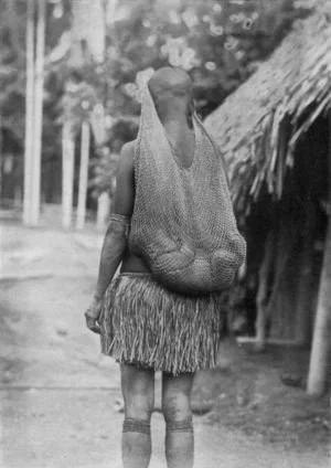 In this archival image, a woman from Papua New Guinea is pictured carrying her baby in a traditional bilum, a handwoven net bag that is an integral part of daily life and identity across the region. The bilum is worn draped over her forehead, with th
