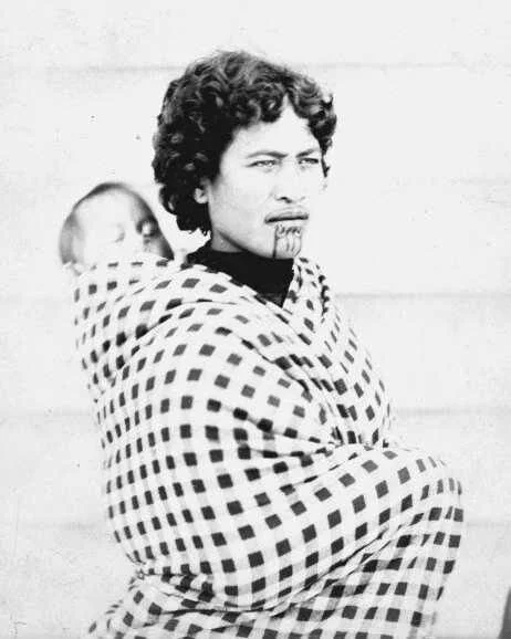 Her gaze is unwavering, her moko kauae etched with mana and meaning. This Māori mother stands proudly with her tamaiti carried snugly on her back, swaddled in a checked woolen cloak that speaks to both practicality and presence. The fabric’s strong g