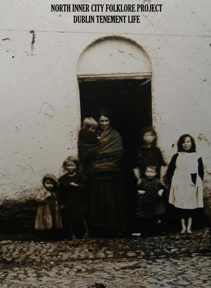 Outside a worn tenement doorway in inner-city Dublin, a mother stands with her children gathered around her. The wall behind them is cracked and whitewashed, the cobbled ground beneath their feet uneven. The children wear layered dresses, aprons, and